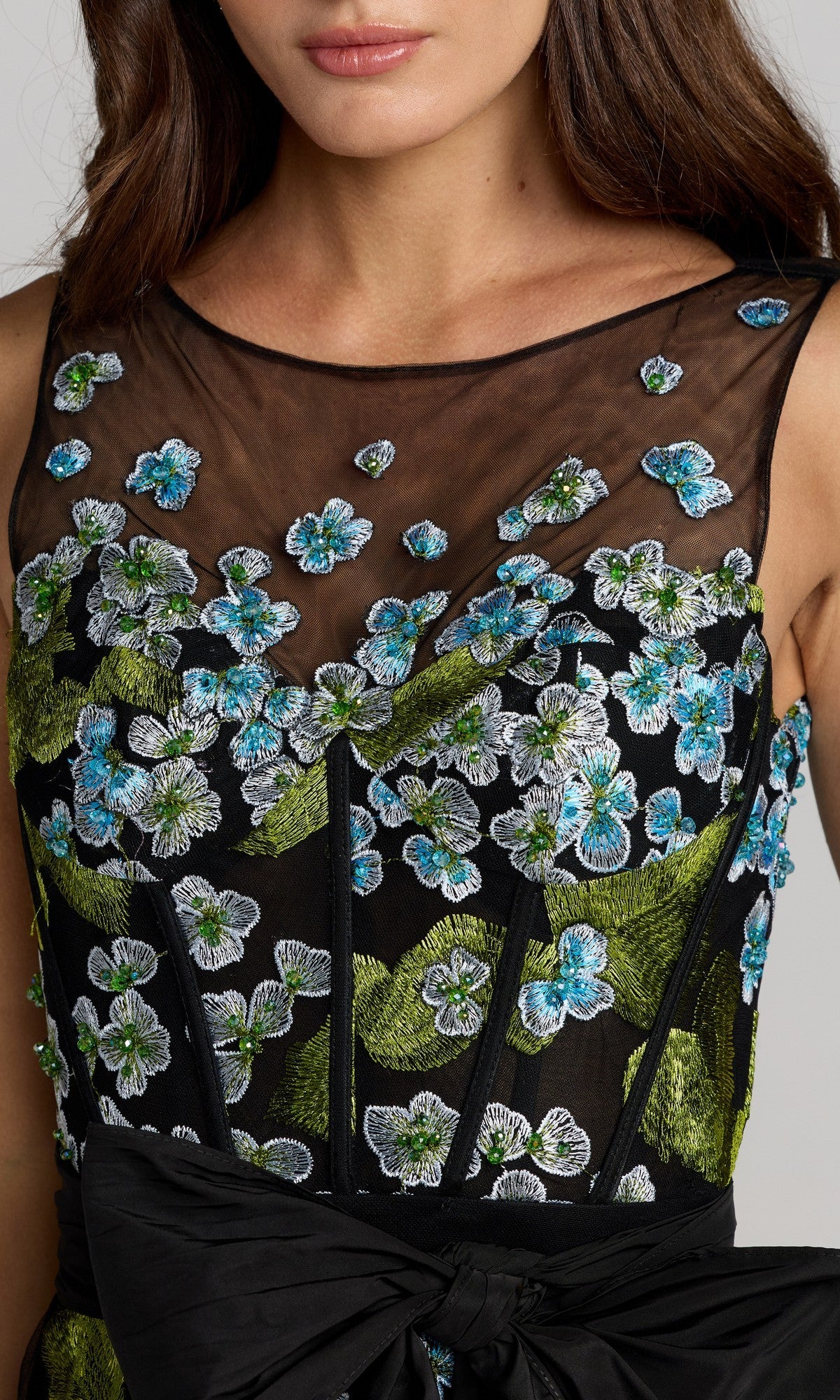 A woman wearing the Mac Duggal 21058 long formal dress, featuring a sleeveless design, sheer mesh, blue, green, and white floral embroidery, and a statement waist bow.